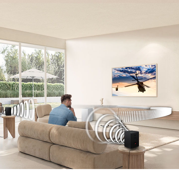 Soundbar Samsung HW-S701D bílý Muž na gauči sleduje televizi, z reproduktorů kolem něj a soundbaru Samsung HW-S701D ležícího pod TV vychází zvuk znázorněný bílými čárami
