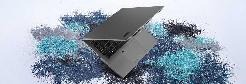 Notebook Acer TravelMate P2 (TMP216-71-G3-TCO-53AR) šedý Elegantní šedý notebook, mírně otevřený, leží na strukturovaném povrchu s rozptýlenými modrými a šedými částicemi.