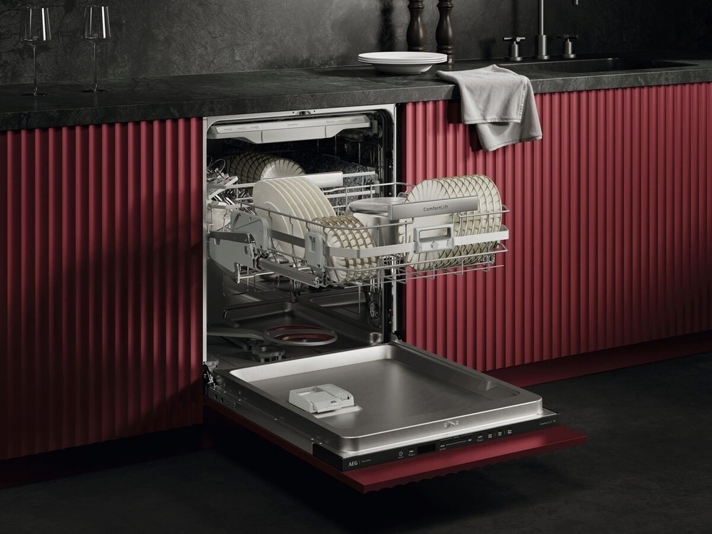 Vestavná myčka AEG NG82IA50TK série 8000 CleaningPerformance otevřená. Spodní koš je díky závěsův ComfortLift v úrovni horního koše a je plně naložen talíři. Myčka je vsazena do kuchyňské linky s červeným dekorem.