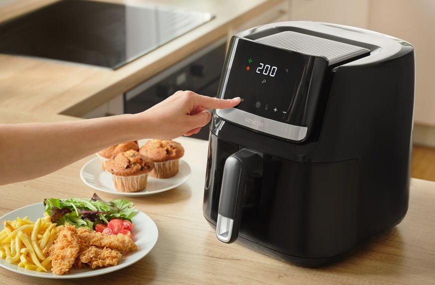 Fritéza horkovzdušná Sencor SFR 6550BK-EUE3 černá Žena nastavuje na LED displeji u horkovzdušné fritézy značky Sencor časovač na 60 minut. Vedle něj se nachází upečené muffiny a stripsy se zeleninou.