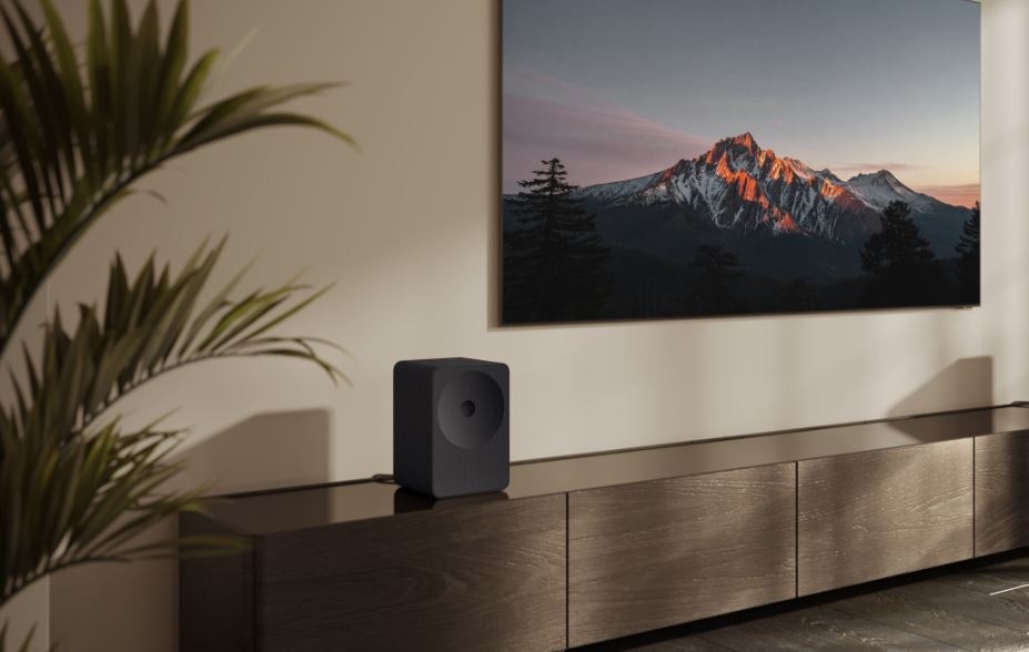 Reproduktor Samsung HW-LS70H Studio 7 pod televizi, která visi na stěně. Samsung HW-LS70H Studio V TV jsou hory a lesy. Reproduktor stojí na televizním stolu, vedle je kytka