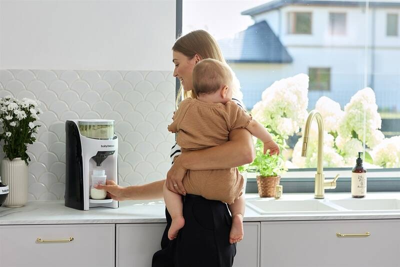 Ohřívač mléka Baby Brezza Formula Pro Advanced Classic bílý Usměvavá maminka drží v jedné ruce miminko a druhou rukou obsluhuje Baby Brezza Formula Pro Advanced Classic.