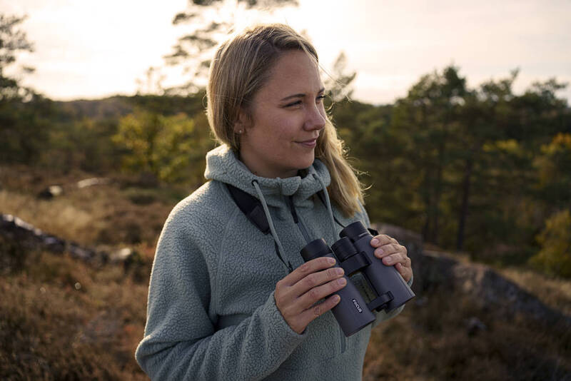 Dalekohled Focus Sports Optics Observer 10x42 ED černý Blondýnka v tyrkysové mikině s kapucí drží šedý dalekohled a rozhlíží se v přírodě při západu slunce.