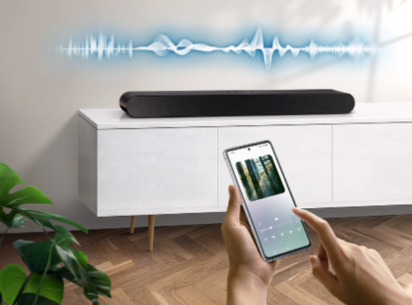 Soundbar Samsung HW-S50B černý Soundbar leží na skříňce v místnosti, nad ním je zakreslena proměnlivá hlasitost zvuku, v popředí ruce drží mobilní telefon s přehrávanou hudbou