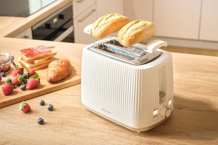 Topinkovač Sencor STS 7510WH bílý Na lince v moderní kuchyni leží topinkovač Sencor STS 7510WH. Vedle něj je na prkénku položený croissant, džem, jahody a borůvky.