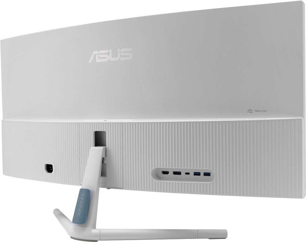 Na obrázku je zadní strana monitoru Asus VU34WCIP-W 34" v bílém provedení z šikmého pohledu. Je vidět široké prohnuté tělo displeje, světlý zadní kryt s jemným svislým žebrováním a decentní logo Asus. Uprostřed je uchycení bílého stojanu se světle modrým detailem. Na zadní straně jsou vidět porty, vlevo napájecí vstup a vpravo panel s obrazovými a USB porty. V pravé horní části je označení Nano-Ion.