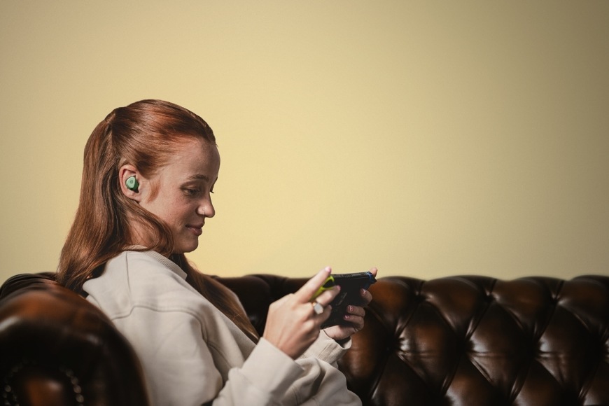 Žena se sluchátkem GameBuds Matcha v uchu drží Nintendo Switch při sezení na pohovce