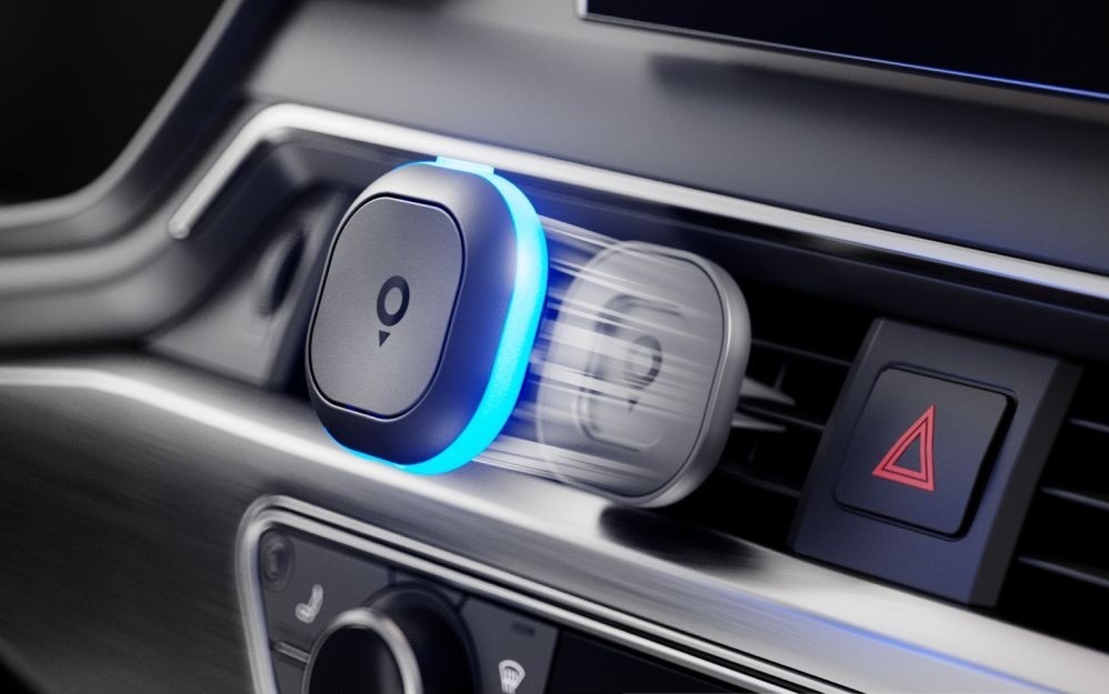Cestovní asistent TomTom Tom připevněný magnetickým držákem k ventilační mřížce automobilu. Modře svítící LED kroužek signalizuje aktivní upozornění a rozmazaný obrys znázorňuje snadné sejmutí zařízení z držáku.