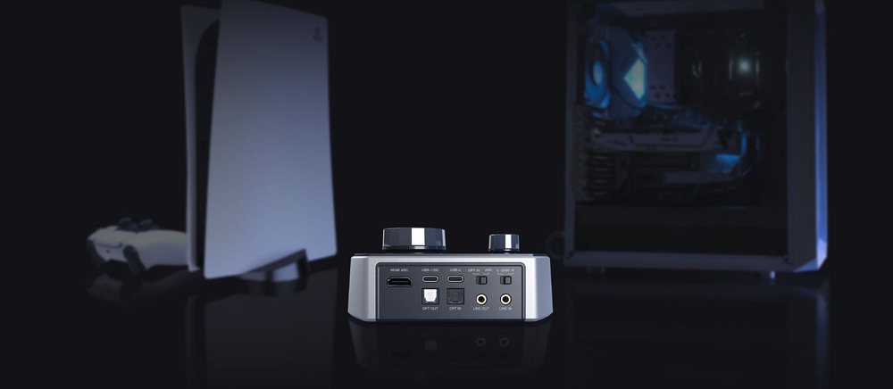Digitální audio převnodník a zesilovač s různými vstupními porty je umístěn na reflexním povrchu. Herní konzole a ovladač jsou rozmazané a umístěné na tmavém pozadí.