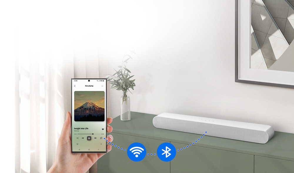 Soundbar Samsung HW-S61D bílý Ruka drží mobilní telefon s přehráváním hudby, na pozadíje soundbar Samsung HW-S61D spojený s mobilem tečkovanou modrou čárou s ikonami se symboly WiFi a Bluetooth