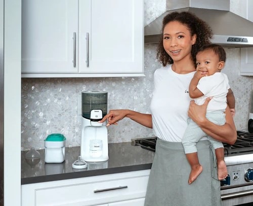 Maminka drží v jedné ruce dítě a druhou rukou chystá vodu do lahvičky z ohřívače vody Baby Brezza Instant Warmer Black.