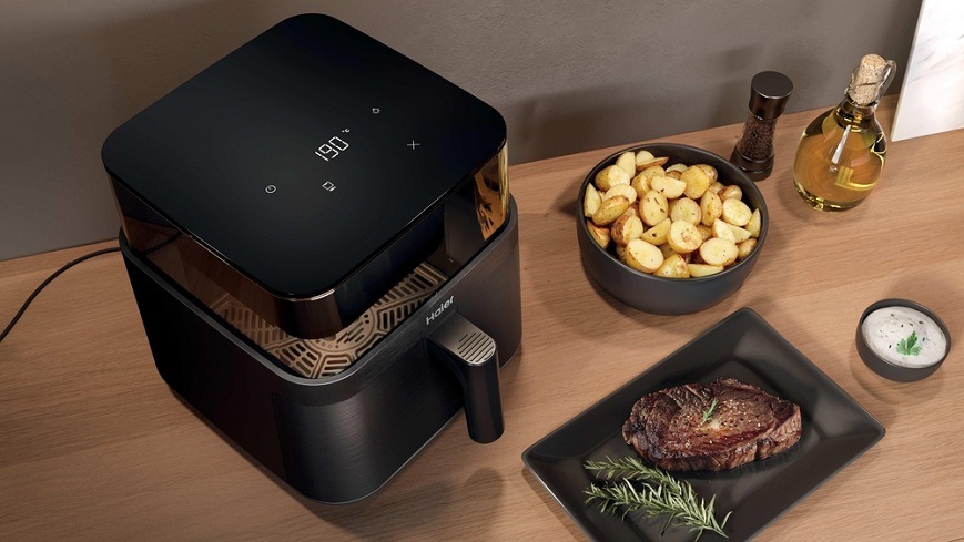 Fritéza horkovzdušná Haier I-Master Series 3 HAF3AFW 011 černá Horkovzdušná fritéza Haier HAF3AFW 011 se zapnutým světlem na dřevěném stole, kolem ní jsou zhotovené pokrmy a dochucovadla