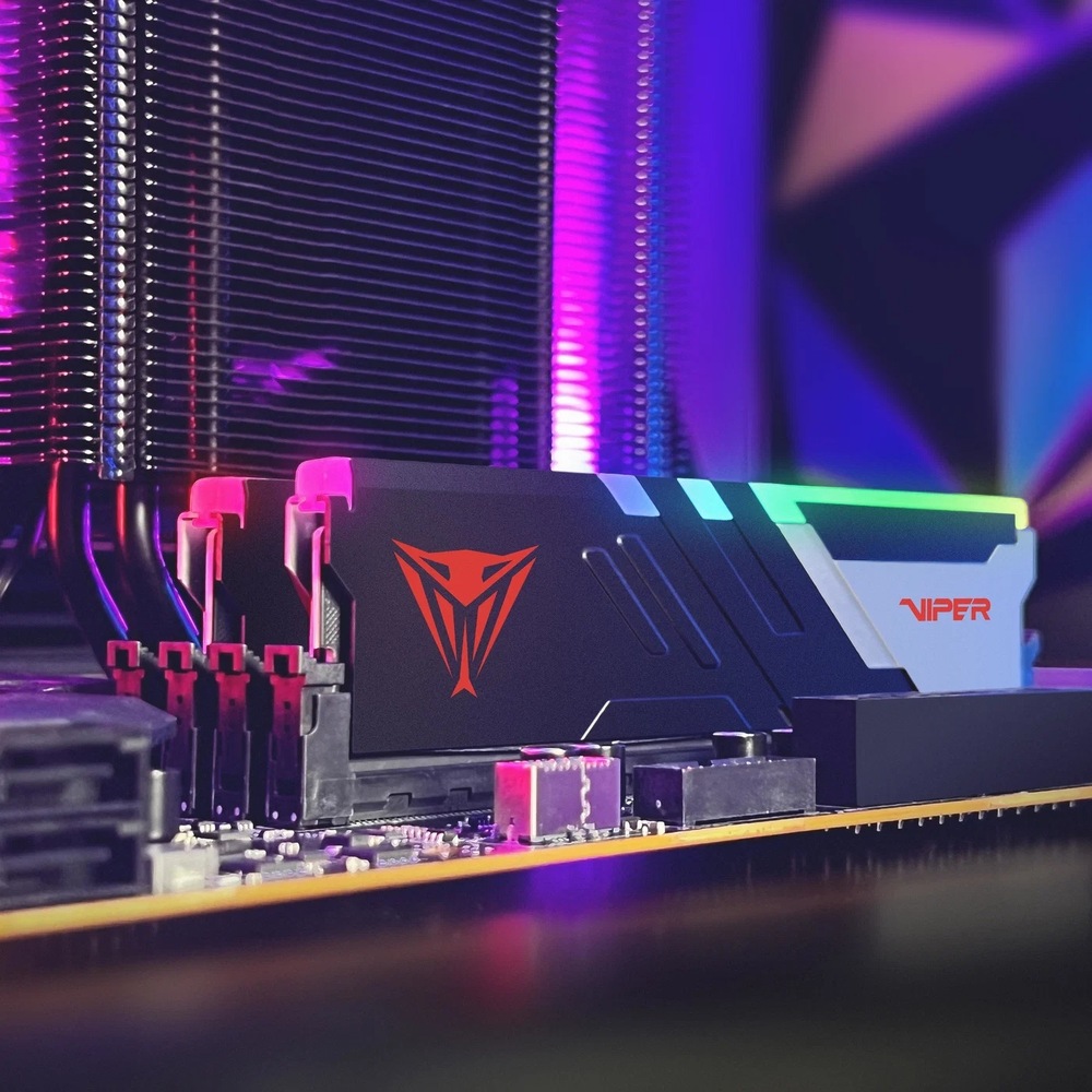 Na obrázku je paměťový modul Patriot Viper Venom RGB osazený v základní desce počítače. Modul má černý chladič se stříbrným detailem, červeným logem Viper a barevně svítící RGB lištou na horní hraně. V pozadí je vidět velký chladič procesoru s kovovými žebry a celé okolí je nasvícené fialovým a růžovým RGB světlem. Snímek ukazuje paměť přímo v sestavené počítačové skříni.
