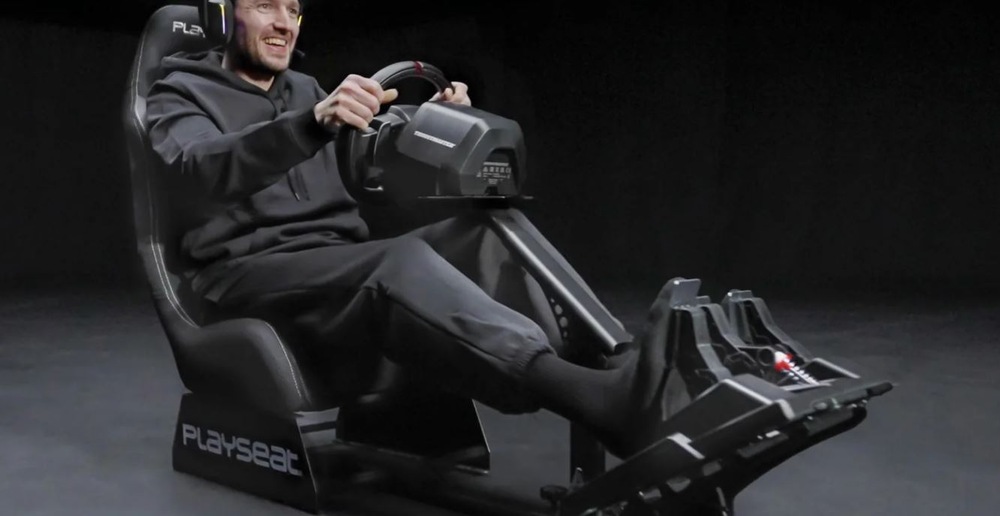 Závodní kokpit Playseat Evolution black ActiFit černý Osoba v černé mikině s kapucí a sluchátky sedí v herním křesle a s úsměvem používá závodní simulátor s volantem a pedály.