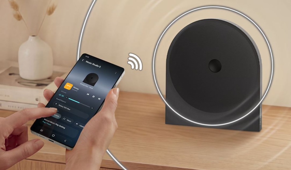 ovladání reproduktoru Samsung HW-LS50H Studio 5 přes mobilni aplikaci v telefonu. Reproduktor je na drevenem stolku, v pozadi je vaza se suchou kvetinou.
