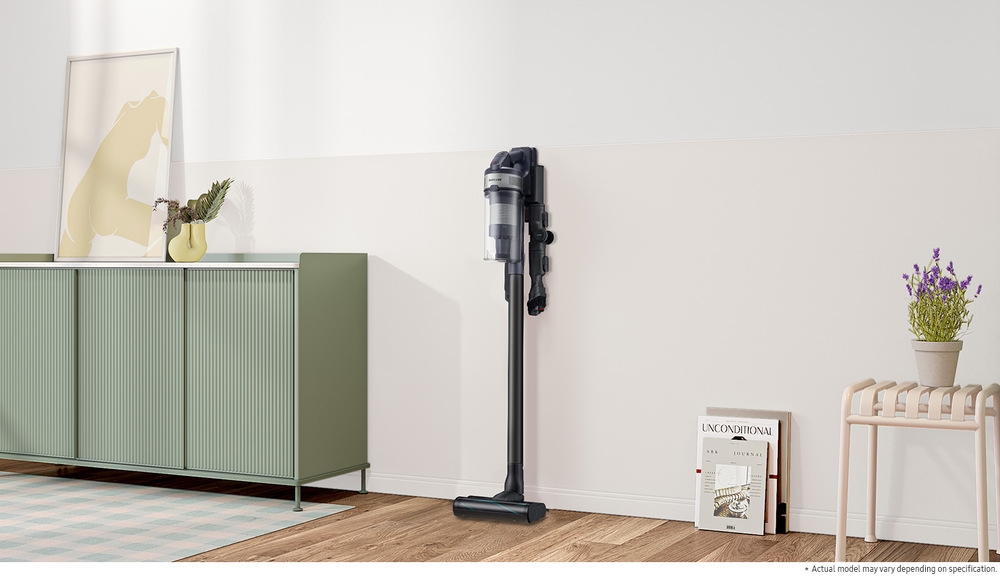Tyčový vysavač Samsung JET 75E complete VS20B75ACR5/GE stříbrný Tyčový vysavač Samsung JET 75E complete VS20B75ACR5/GE opřený o zeď místnosti, vlevo je skříňka, vpravo obraz opřený o zeď a stolek s květinou v květináči