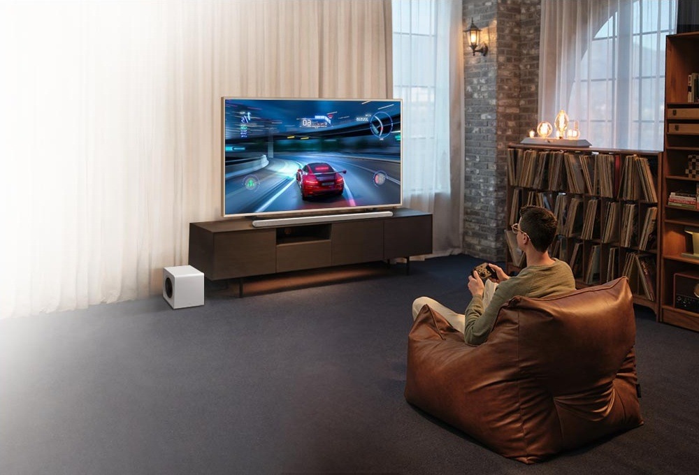 Samsung HW-S801D leží na televizním stolku před televizí, v popředí sedí muž na sedacím vaku a hraje na televizi videohru