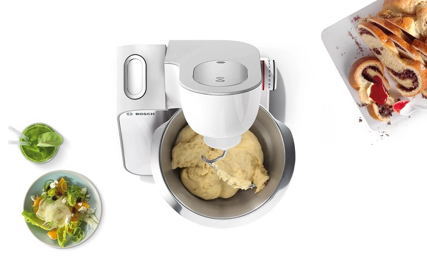 Kuchyňský robot Bosch MUM5 MUM58251 bílý Pohled shora na kuchyňský robot Bosch MUM 5, který má zvednuté rameno a v míse těsto. Vedle robota je upečený závin a miska se salátem