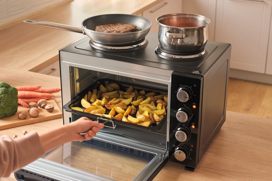 Mini trouba Sencor SEO 2800BK s otevřenými dvířky při pečení brambor, na horní varné ploše se připravuje steak na pánvi a omáčka v hrnci v moderní domácí kuchyni.
