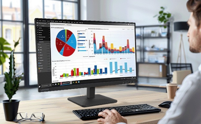 Monitor Lenovo L27-4e černý Osoba v kanceláři si prohlíží datové grafy na širokoúhlém monitoru, zobrazující barevné grafy a koláčové grafy. Mezi předměty na stole patří klávesnice, hrnek a brýle.