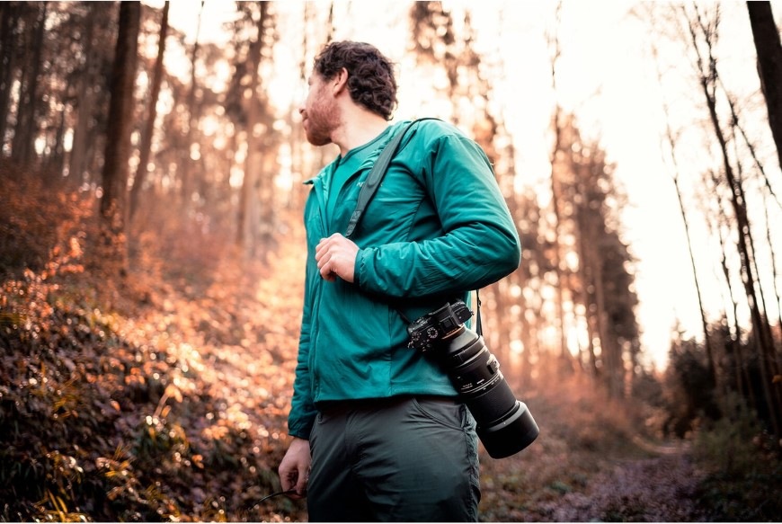 Muž v lese nesoucí fotoaparát s objektivem Tamron 150–500 mm f/5–6.7