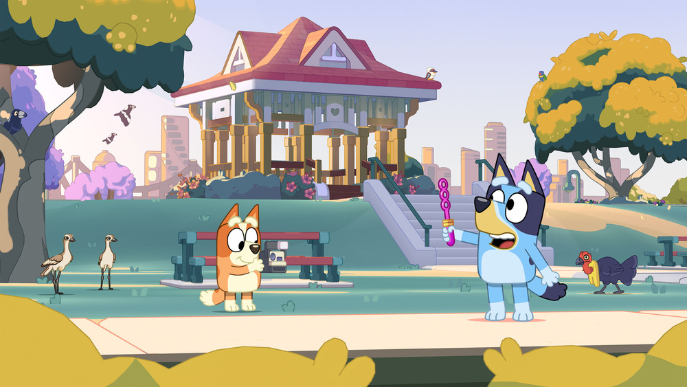 Bluey a Bingo stojí ve slunném parku s altánem a stromy. Bluey drží bublifuk. V pozadí jsou ptáci a slepice, což celé scéně dodává hravou atmosféru.