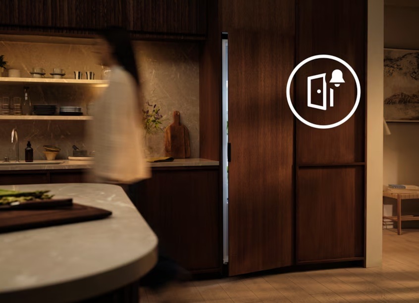 Pootevřená vestavná lednice Electrolux. Na skříni vedle je symbol alarmu. Před lednicí je zachycena procházející žena.