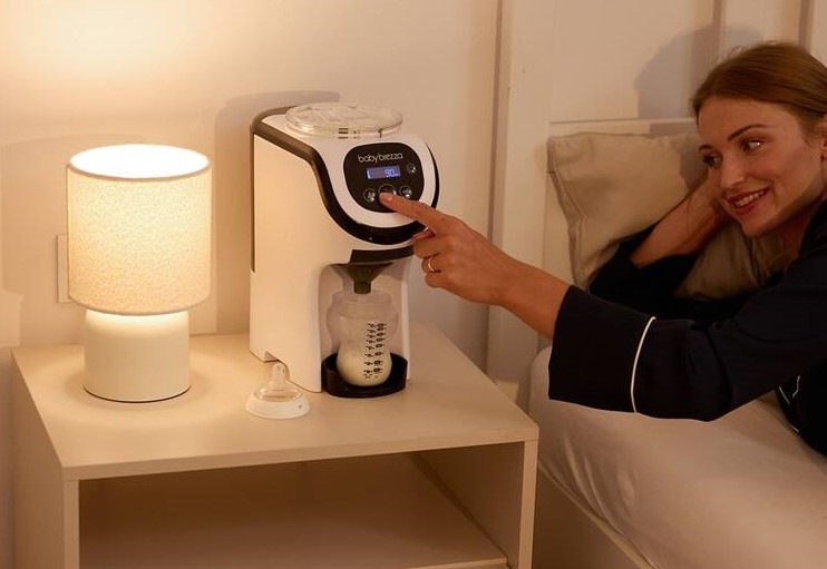 Ohřívač mléka Baby Brezza Formula Pro Mini je postavený na nočním stolku vedle lampičky, maminka přímo z postele chystá krmení.