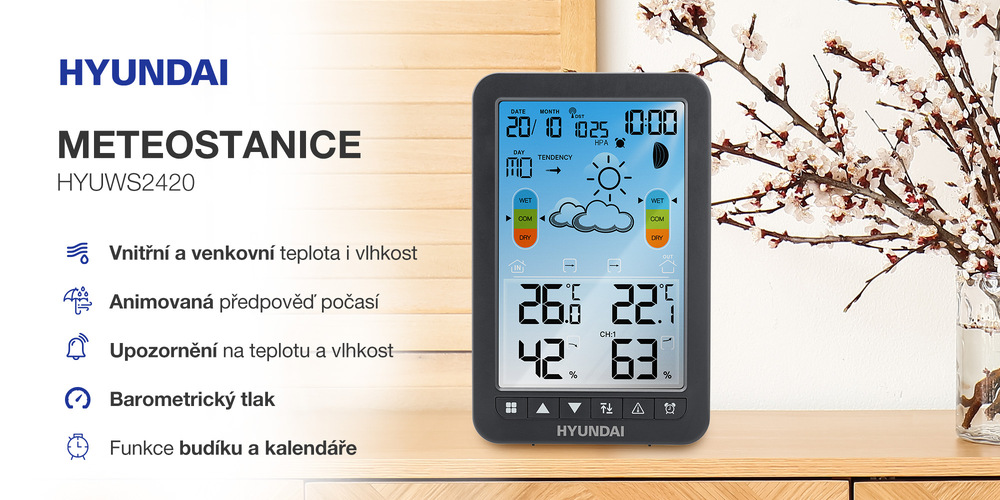 Černá meteostanice Hyundai HYUWS2420 zobrazená v interiéru vedle rozkvetlé větvičky. Displej ukazuje vnitřní a venkovní teplotu, vlhkost, barometrický tlak, čas a kalendář