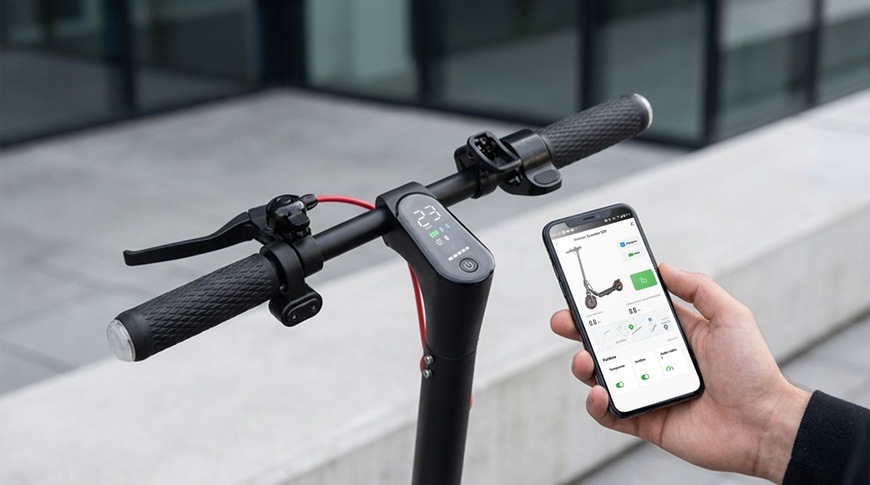 Detail řídítek elektrické koloběžky Sencor SCOOTER S31 s integrovaným displejem, který zobrazuje aktuální rychlost a další údaje. Ruka vedle drží chytrý telefon s aplikací Sencor HOME pro ovládání koloběžky a sledování jejích funkcí.