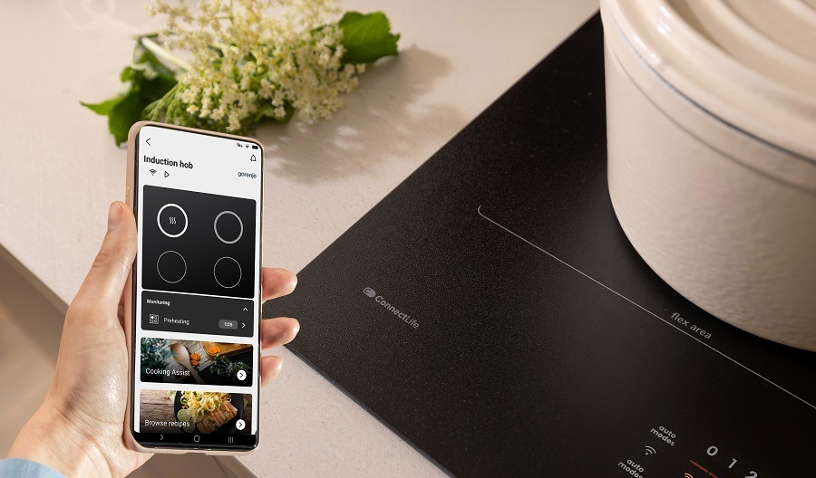 Indukční varná deska Gorenje G600 GI6442MSCWF InfiniteMatt černá ruka drží mobilni telefon u varne desky gorenje, se kterou je možno kounikovat. V telefonu je zapnuta aplikace ConnectLife.