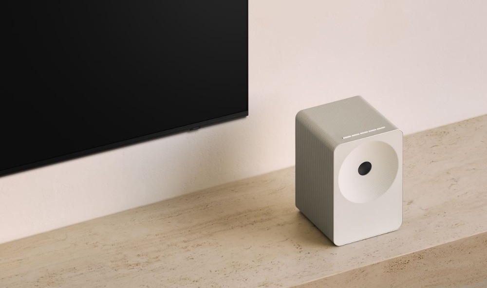 Reproduktor Samsung Music Studio 7 LS71H (1 ks) bílý Bílý reproduktor Samsung HW-LS71H Studio 7 stojí na dřevěném stolku pod vypnutou televizi, která je zavěsena na zdi.