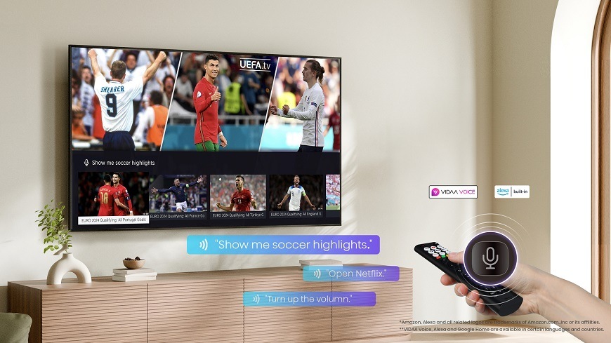 Na obrázku je televize Hisense 50E7PRO umístěná v moderním obývacím pokoji, na jejíž obrazovce běží sportovní přenos s fotbalovými hráči. V popředí drží uživatel dálkový ovladač s aktivovaným mikrofonem, který umožňuje hlasové ovládání televizoru – například vyhledání sportovních sestřihů, otevření Netflixu nebo úpravu hlasitosti.