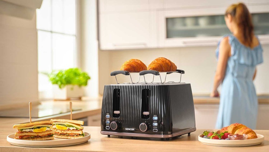 Topinkovač Sencor STS 7541BK černý Designový topinkovač Sencor opéká croissanty, vedle je připravený sendvič a ovoce na snídani.