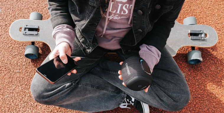 Mladý člověk drží reproduktor Concept SoundJoy BR5010 a telefon při odpočinku u longboardu