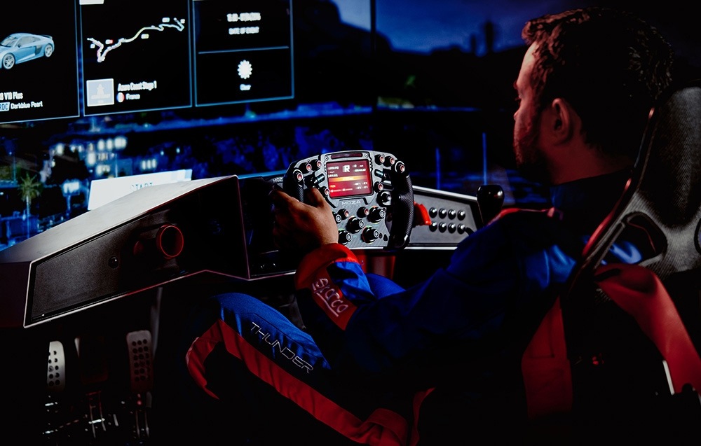 Základna Moza Racing R25 Ultra Direct Drive hliník Osoba v závodní kombinéze sedí v řidičském simulátoru a drží volant. Na několika obrazovkách se zobrazují závodní data a mapy tratí, což vytváří soustředěnou a pohlcující atmosféru.