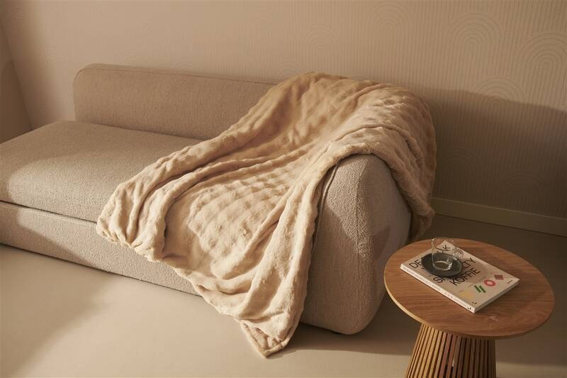 Vyhřívací deka Duux DXOB11 Yentl Cuddle 200 x 200 cm béžová Nadrozměrná béžová deka Duux Yentl volně přehozená přes textilní pohovku vedle dřevěného stolku s časopisem v jemně laděném moderním obývacím prostoru.