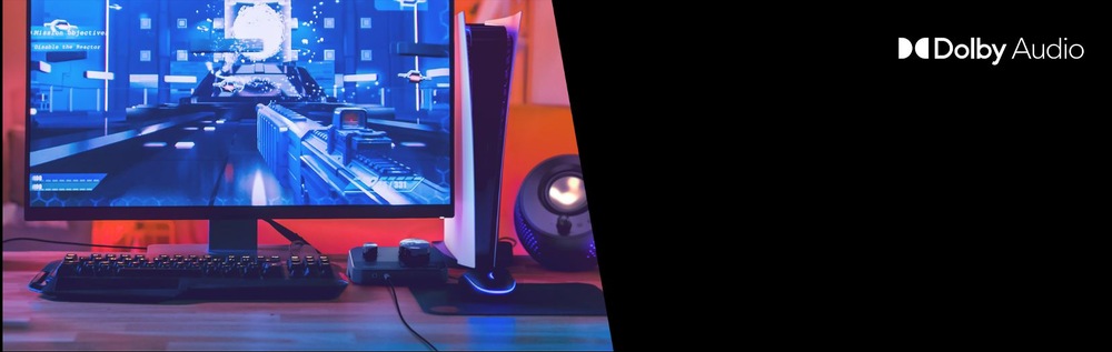 Herní sestava s monitorem, konzolí a reproduktory na stole. Na obrazovce je zobrazena futuristická hra. Vpravo je logo Dolby Audio. Atmosféra působí hi-tech a pohlcujícím dojmem.