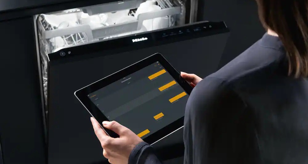 Myčka nádobí Miele G 7040 SC AutoDos nerez Žena drží v rukou tablet, na pozadí je vestavná myčka MIele s otevřenými dvířky
