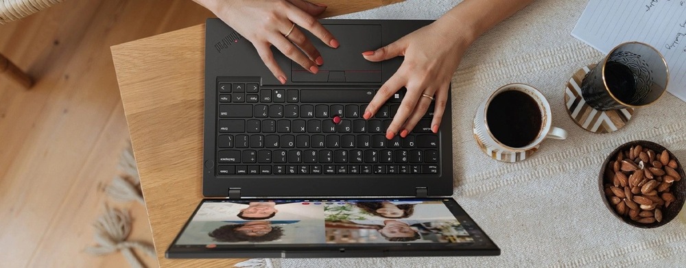 Notebook Lenovo ThinkPad L14 Gen 6 černý Uživatel píše na notebooku, na kterém je vidět videohovor se čtyřmi lidmi. Vedle něj stojí šálek černé kávy, mandle a zápisník, které vytvářejí útulnou a produktivní atmosféru.