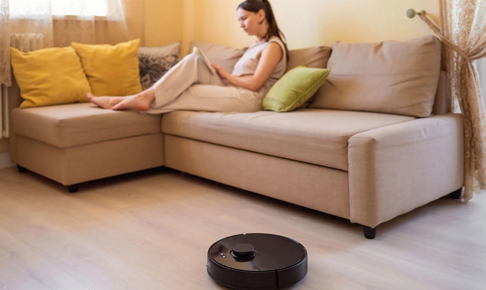 žena sedí na sedačce, čte si knížku a kolem projíždí robotický vysavač Dreame F20 Plus v černé barvě.