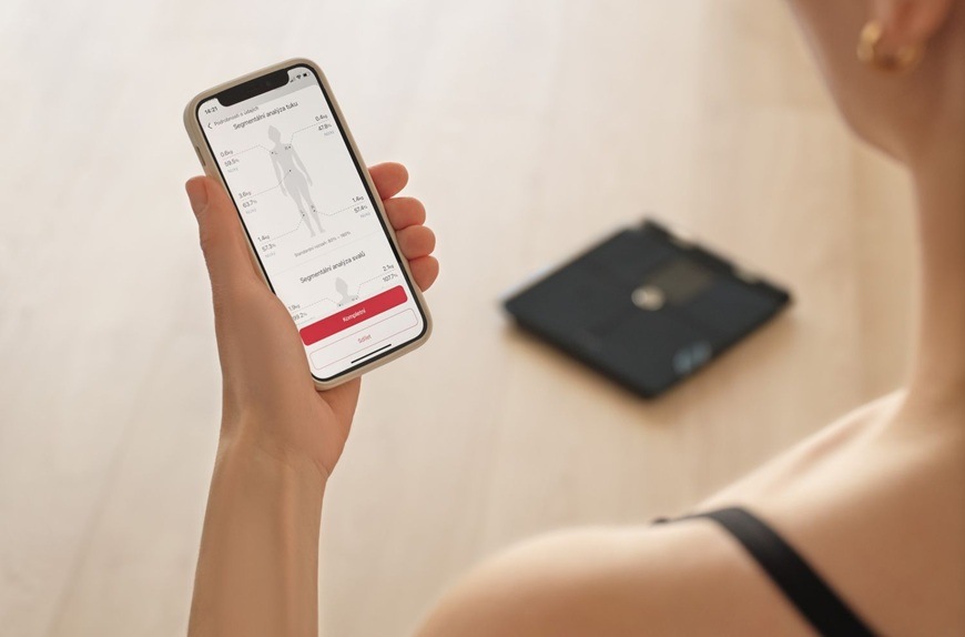 Osobní váha Sencor SBS 9102BK modrá Žena drží v ruce chytrý telefon s aplikací zobrazující analýzu těla a měření tělesných hodnot. V pozadí na podlaze leží chytrá osobní váha Sencor SBS 9102BK. Celá scéna působí čistě a moderně a zdůrazňuje propojení váhy s mobilní aplikací.