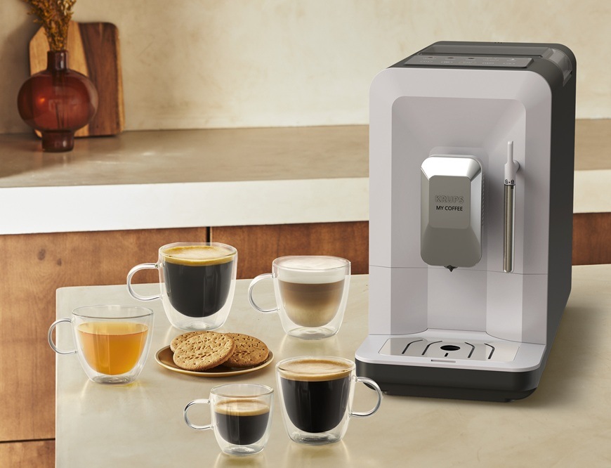 Automatický kávovar Krups EA201BE0 My Coffee Steam je položen na kuchyňské lince. Vedle něj jsou skleněné šálky s různými kávami a čajem. Na talířku jsou sušenky. V pozadí je vidět zbytek kuchyně.