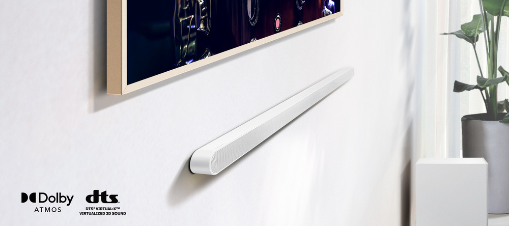 Soundbar Samsung HW-S701D bílý Bílý soundbar Samsung HW-S701D je připevněný na zdi pod televizí Samsung Frame, vpravo je rostlina v květináči, v levém dolním rohu jsou loga Dolby ATMOS a DTS