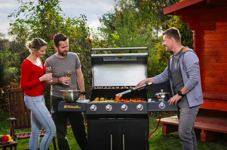 Tři lidé připravují maso a zeleninu na plynovém grilu během zahradního posezení