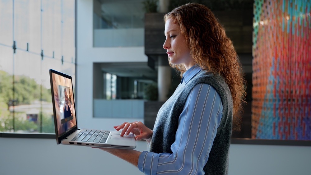 Žena s kudrnatými vlasy drží notebook a účastní se videohovoru. Stojí v moderní profesionálně vypadající kanceláři s velkými okny a barevnými uměleckými díly.