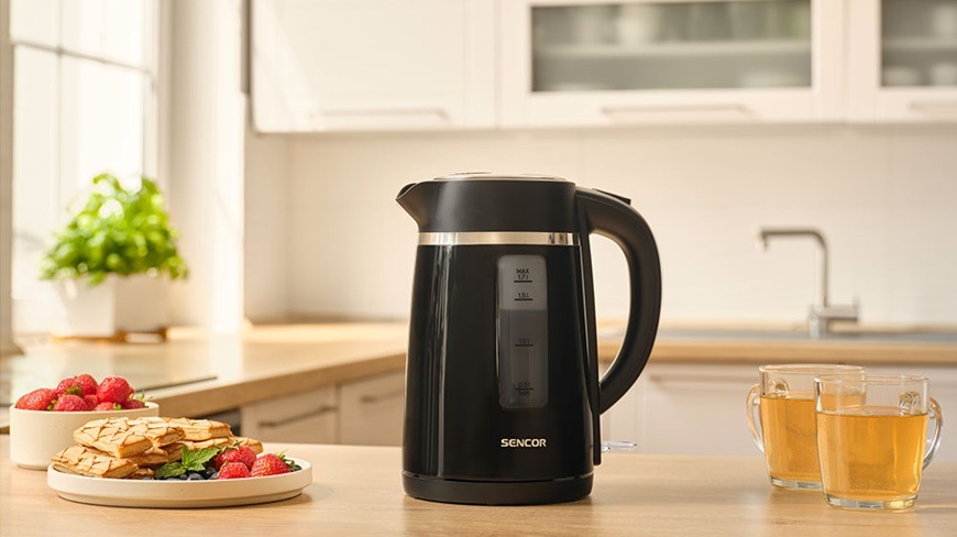 Rychlovarná konvice Sencor SWK 2710BK, 1,7 l černá Rychlovarná konvice Sencor SWK 2710BK je položená na lince. Vedle je položená mísa s jahodami, talíř se snídaní a na druhé straně jsou dva šálky s čajem.