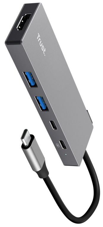 Na obrázku je stříbrný USB-C hub Trust DALYX 5v1 zachycený šikmo zepředu. Má úzké kovové tělo s jednoduchým hranatým tvarem a z jednoho konce z něj vede krátký kabel zakončený USB-C konektorem. Na boku jsou dva modře označené USB porty, na čele HDMI konektor a na další straně dva menší USB-C porty.
