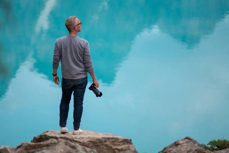 Muž stojící u jezera s odrazem hor drží v ruce fotoaparát Nikon s objektivem NIKKOR Z 24–120 mm f/4 S