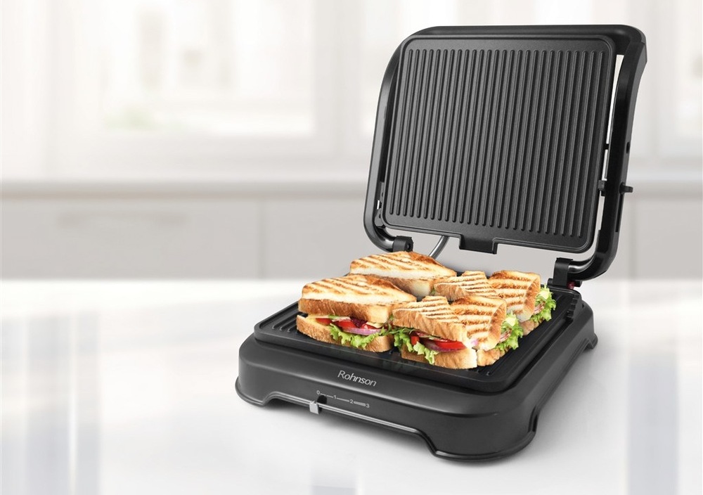 Kontaktní gril Rohnson R-2344 stojí na kuchyňském stole, gril je otevřený a na pečicí ploše leží grilované toasty.